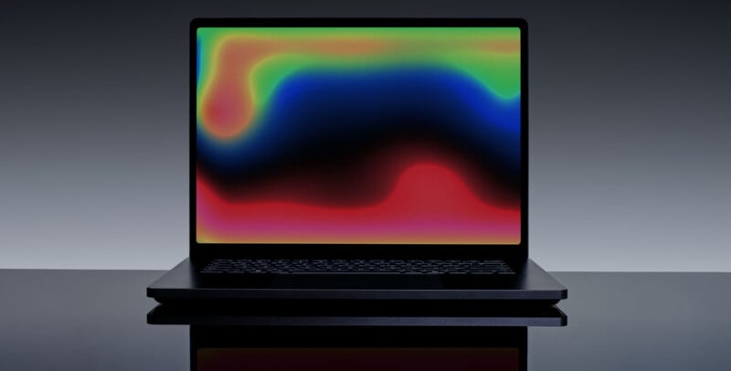 A black laptop on a reflective surface displays a colorful abstract pattern with blended red, blue, green, and yellow shapes on its screen against a gray background.