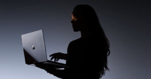 A person with long hair is silhouetted against a dark background, holding and using a laptop with a visible Windows logo. Light highlights part of their face and hands.