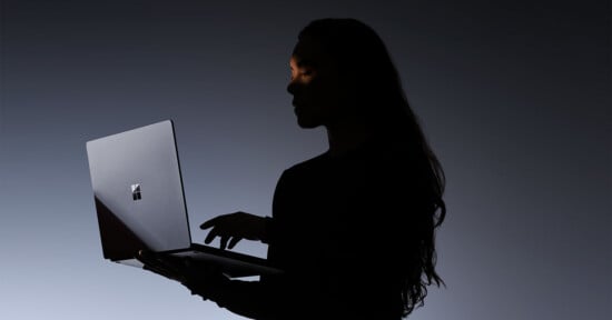 A person with long hair is silhouetted against a dark background, holding and using a laptop with a visible Windows logo. Light highlights part of their face and hands.