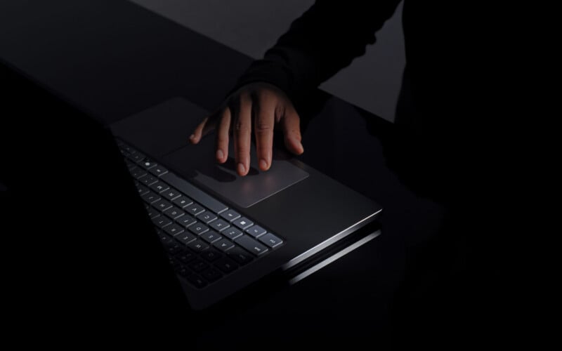 A person’s hand rests on the trackpad of a laptop, illuminated by dim light, with the surrounding area and background mostly in shadow.