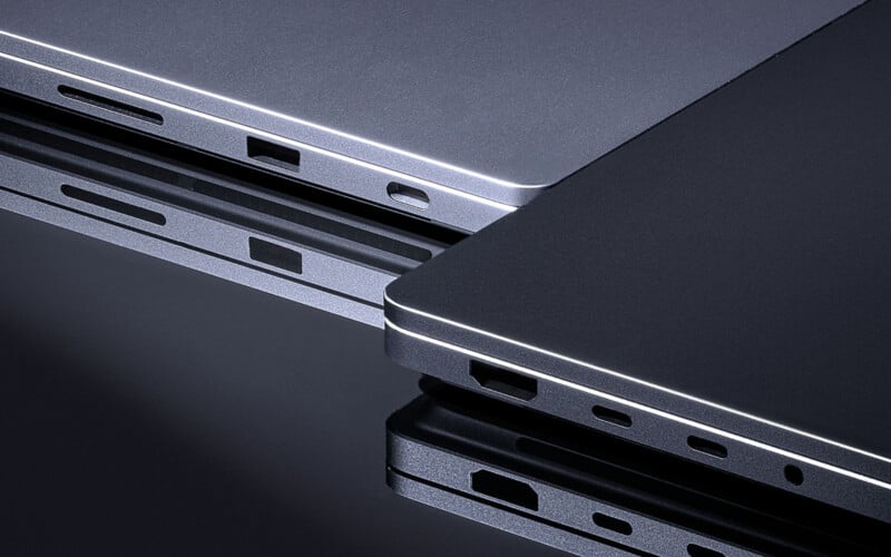 Two sleek, silver and black laptop edges are placed side by side on a reflective surface, showing their various ports and slim profiles. The lighting highlights the metallic finish and modern, minimalist design.