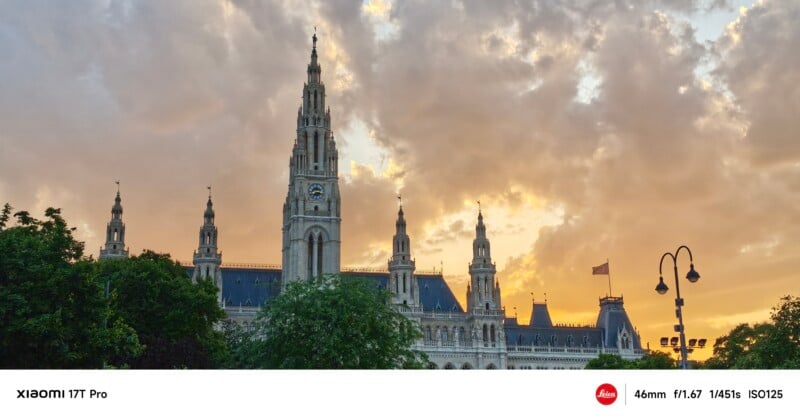 Gothic-style building with tall spires and a clock tower, photographed at sunset with dramatic, orange-clouded sky and trees in the foreground. Street lamps are visible on the right.