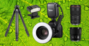 A digital camera with a ring flash, a tripod, a flash, and two camera lenses are arranged on top of a vibrant green leaf with visible veins and water droplets.