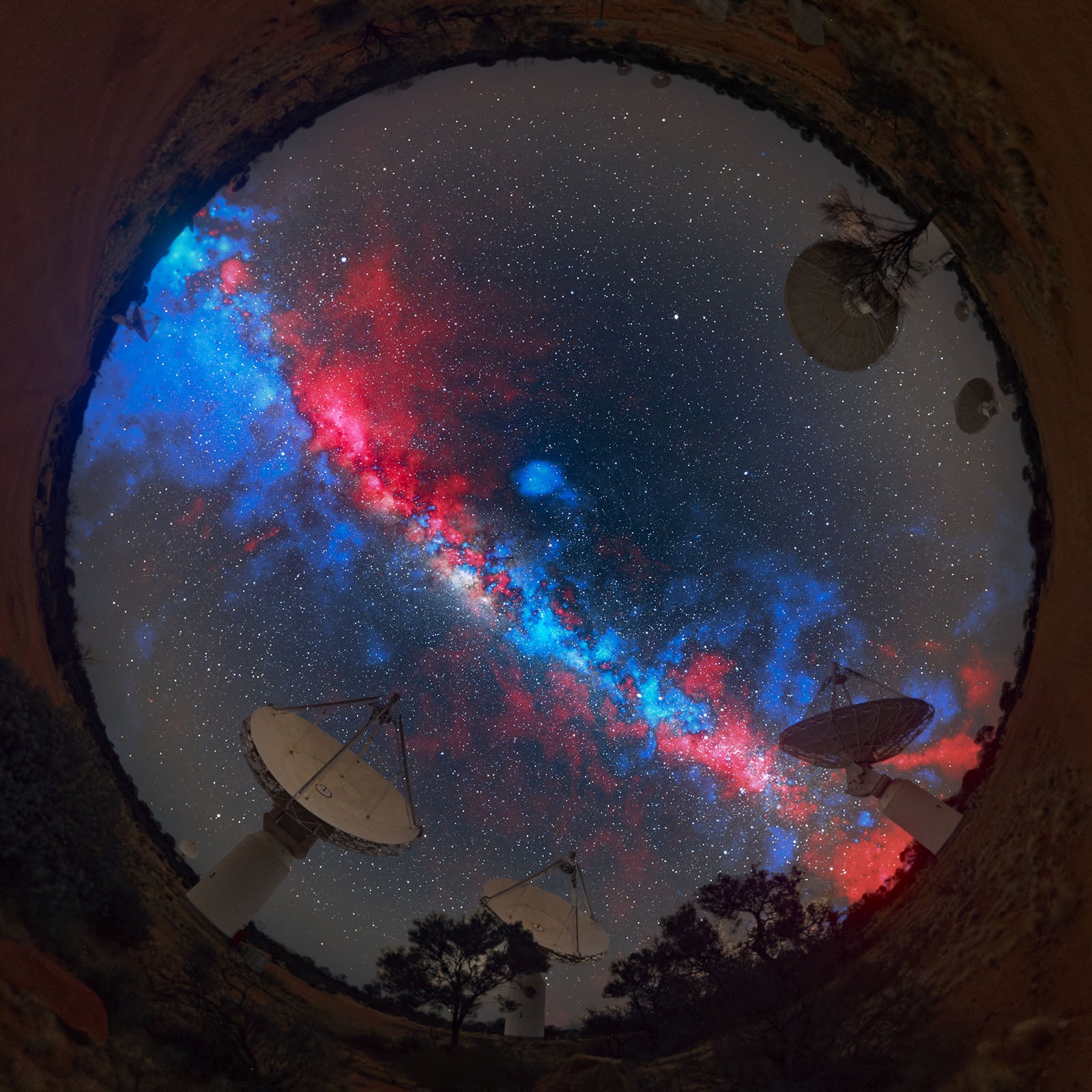 A circular fisheye view of radio telescopes under a vivid, colorful Milky Way night sky, with red, blue, and white hues stretching across the stars above the satellite dishes and silhouetted trees.