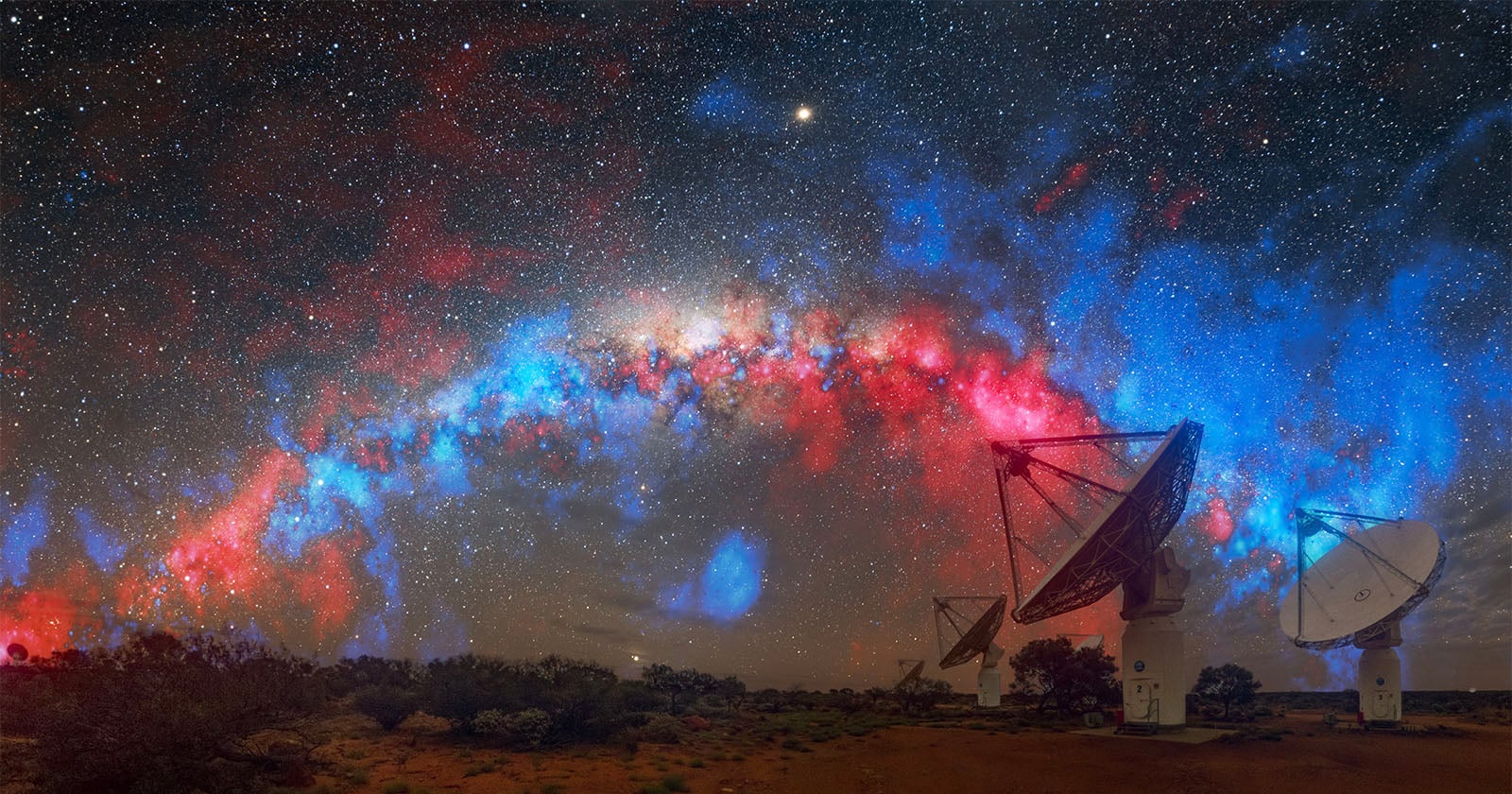 Large radio telescopes stand beneath a vivid night sky filled with stars and a colorful arc of red and blue nebulae, part of the Milky Way galaxy, stretching across the horizon.