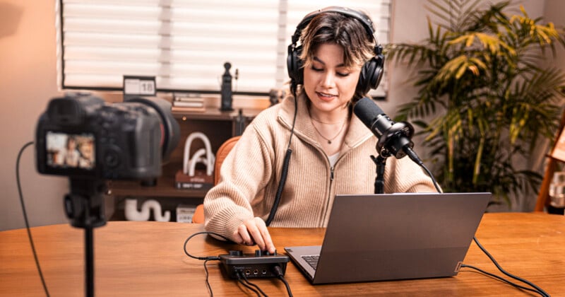 A woman wearing headphones speaks into a microphone while adjusting audio controls, sitting at a desk with a laptop. A camera records her, and there are plants and books in the background.