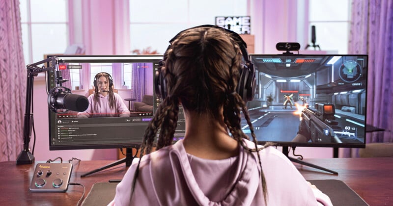 A person with headphones and braided hair streams a video game from a desktop setup with two monitors, one showing their live webcam feed and chat, and the other displaying a first-person shooter game.