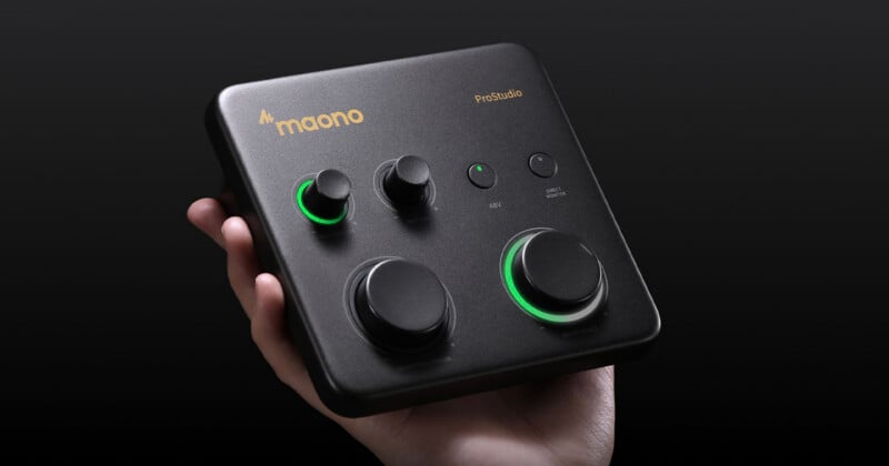 A hand holds a black Maono PreStudio audio interface with multiple knobs, buttons, and two green-lit dials, set against a dark background.