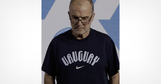 A man wearing glasses and a navy blue t-shirt with the word "Uruguay" and a Nike logo stands looking down in front of a blue and white geometric background.