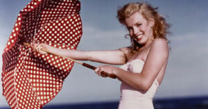 A smiling woman in a light-colored swimsuit holds a red and white polka dot umbrella at the beach, with blue sky and water in the background.