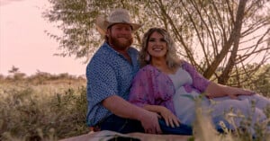 A couple sits together outdoors on a blanket, smiling at the camera. The man wears a cowboy hat and patterned shirt, while the woman wears a flowing pink and white dress. Trees and grass surround them in soft sunlight.