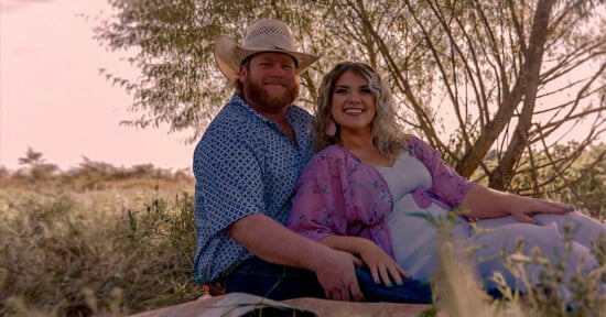 A couple sits together outdoors on a blanket, smiling at the camera. The man wears a cowboy hat and patterned shirt, while the woman wears a flowing pink and white dress. Trees and grass surround them in soft sunlight.