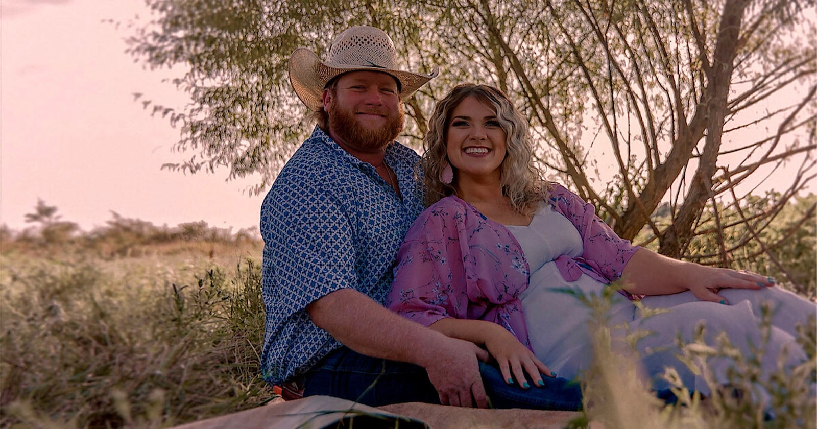 A couple sits together outdoors on a blanket, smiling at the camera. The man wears a cowboy hat and patterned shirt, while the woman wears a flowing pink and white dress. Trees and grass surround them in soft sunlight.