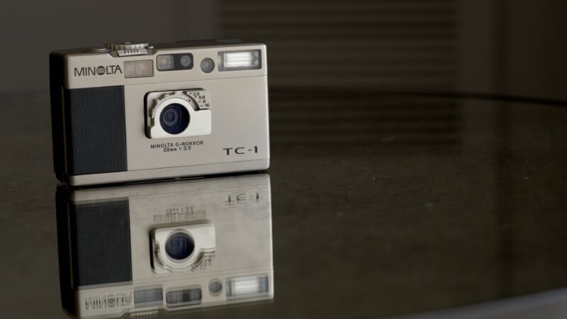 A silver Minolta TC-1 camera sits on a reflective glass surface, creating a clear mirror image beneath it. The background is softly blurred.