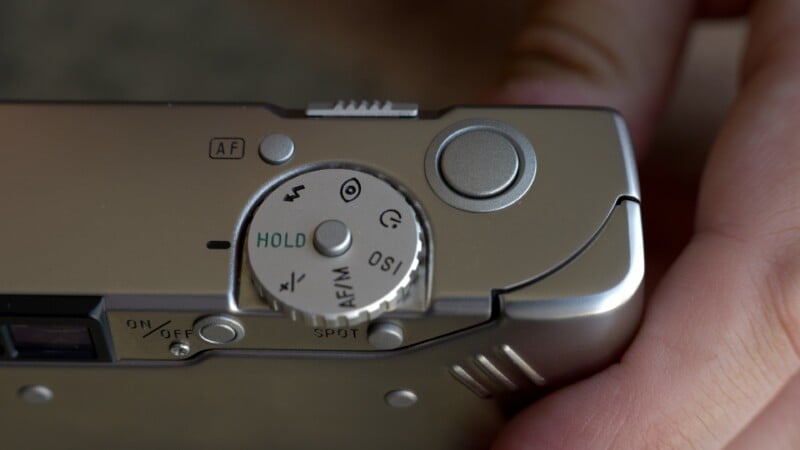 Close-up of a hand holding a camera, focusing on the mode dial and buttons on the top of the camera body. The dial displays various settings, including "HOLD," "ISO," and shooting modes.