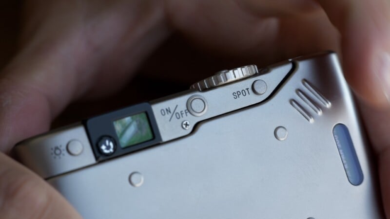 Close-up of a person’s hand holding a silver camera, showing the ON/OFF switch, a SPOT button, control dial, and a small viewfinder on the top surface.