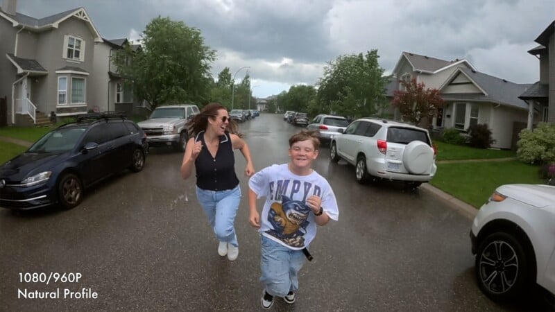A woman and a boy joyfully run down a wet residential street lined with cars and houses on a cloudy day. Both are smiling, wearing casual clothes and sneakers. Text in the corner reads "1080/960P Natural Profile.