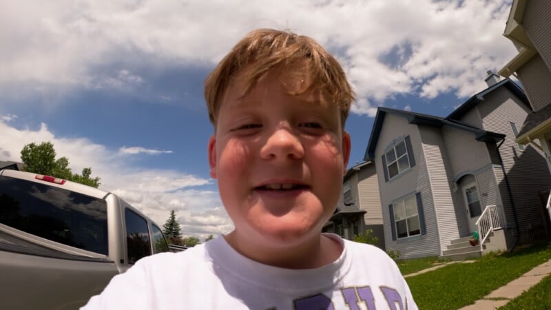 A smiling boy with light brown hair stands outside on a sunny day, wearing a white shirt. Behind him are houses, a green lawn, a blue sky with clouds, and part of a silver vehicle.