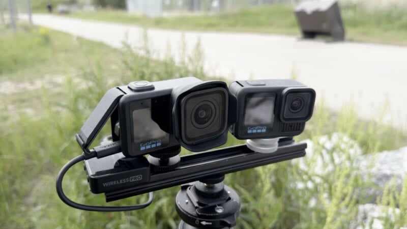 Three GoPro cameras are mounted on a tripod outdoors, surrounded by grass, with a blurred path and objects in the background. The setup appears to be for filming or photography.
