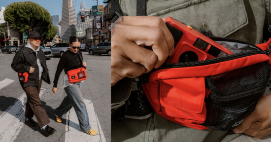 Two people wearing sunglasses walk across a city street, each carrying bright red crossbody bags. A close-up shows a hand placing a red camera into the bag's zippered pocket. Tall buildings and trees are visible in the background.