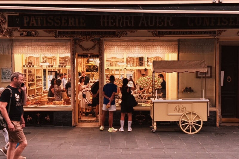 People stand outside and inside a charming French patisserie with large windows, ornate signage, and a yellow cart on the sidewalk. The shop is warmly lit and displays pastries and treats.