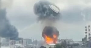 A large fire and thick black smoke billow from a building in an industrial area during daytime, with flames visible at the base and dark clouds rising into the sky.