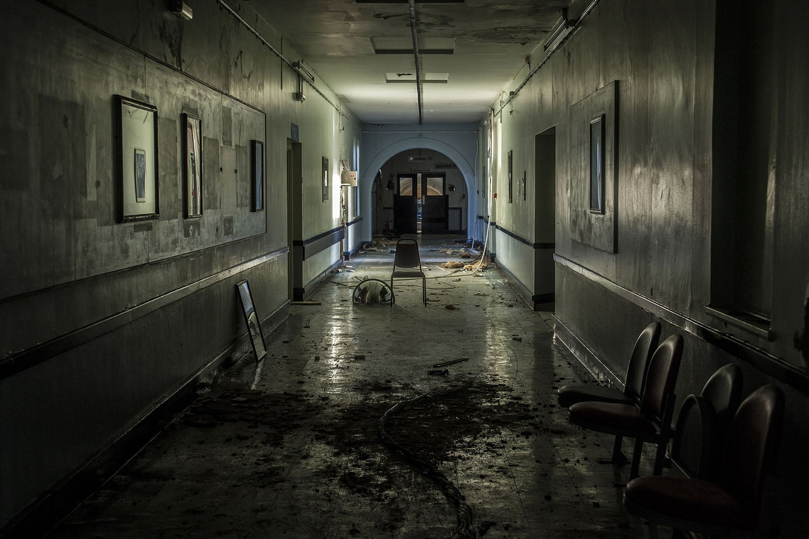 A dimly lit, abandoned hallway with peeling paint, debris on the floor, empty chairs, and scattered objects, creating a sense of neglect and decay. Light filters in from a distant doorway.