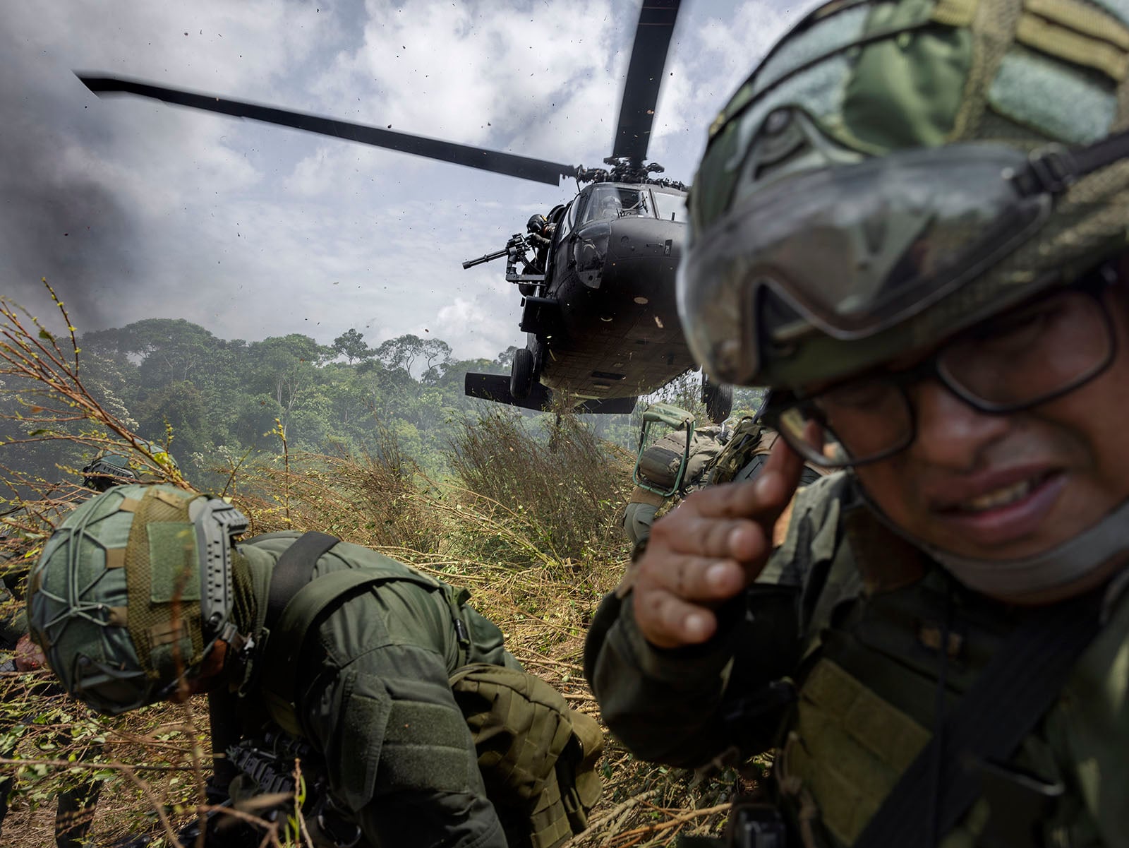 Soldiers in camouflage gear take cover as a military helicopter hovers low to the ground, kicking up dirt and debris in a grassy field with trees and cloudy skies in the background.
