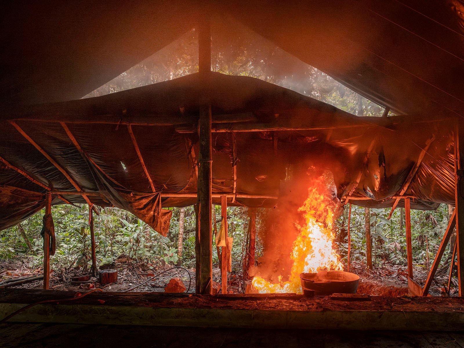 A fire burns intensely inside a rustic, open-sided tent in a forested area, with flames rising from a large container under a makeshift roof, surrounded by trees and dense vegetation.