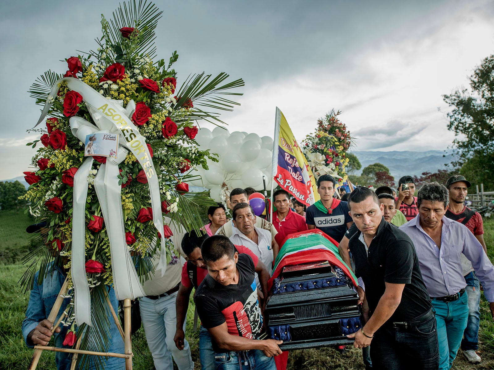 A group of people carry a colorful casket during a funeral procession outdoors, surrounded by floral wreaths, balloons, and a cloudy sky in the background. Mourners walk closely together in a rural setting.