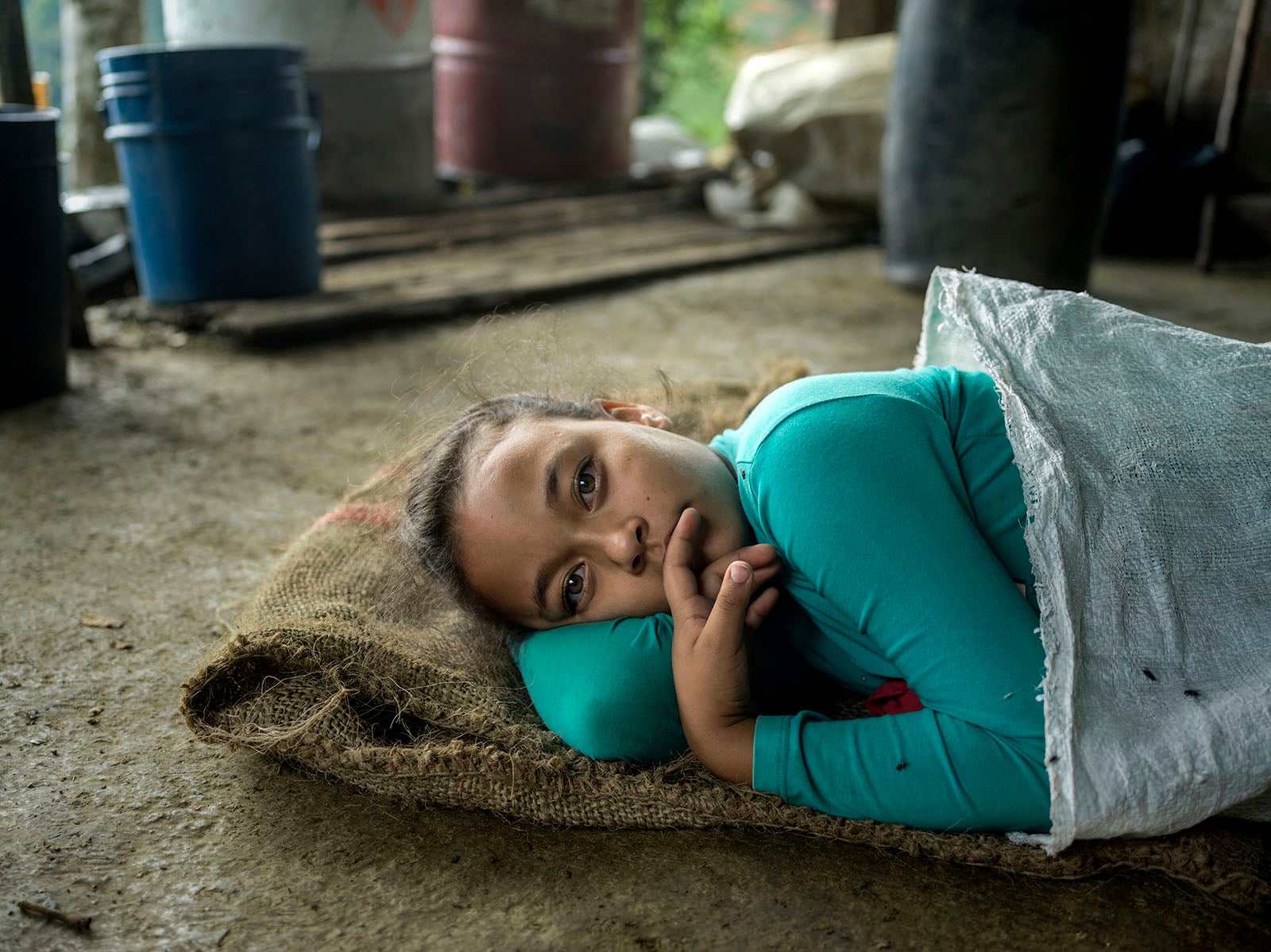A young girl in a turquoise shirt lies on a burlap sack on the floor, partially covered with a white sheet, looking thoughtfully at the camera. The background shows plastic barrels and a rustic setting.