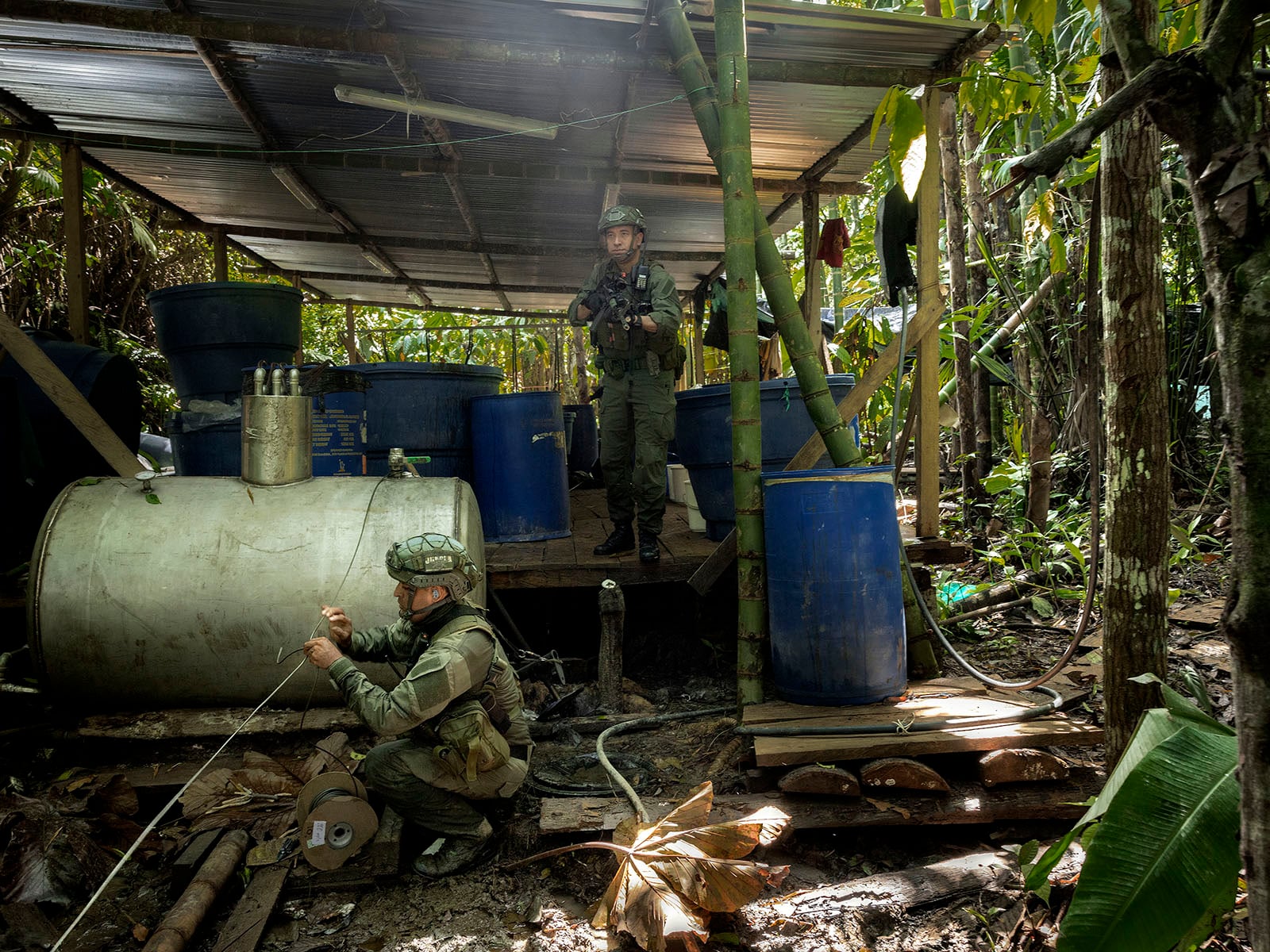 Two soldiers in camouflage gear inspect makeshift equipment and blue barrels under a metal roof in a dense jungle, possibly investigating an illegal operation. One stands guard while the other examines wiring near a large metal tank.