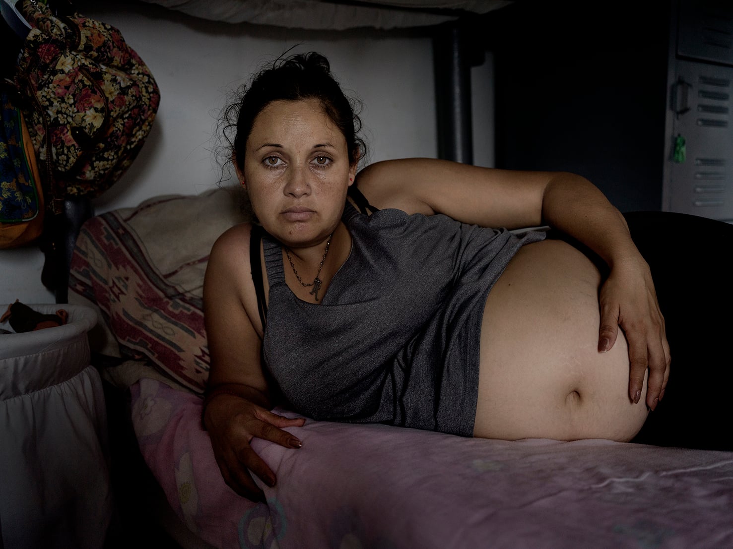 A pregnant woman lies on her side on a bed in a dimly lit room, resting one hand on her exposed belly and looking directly at the camera with a serious expression.