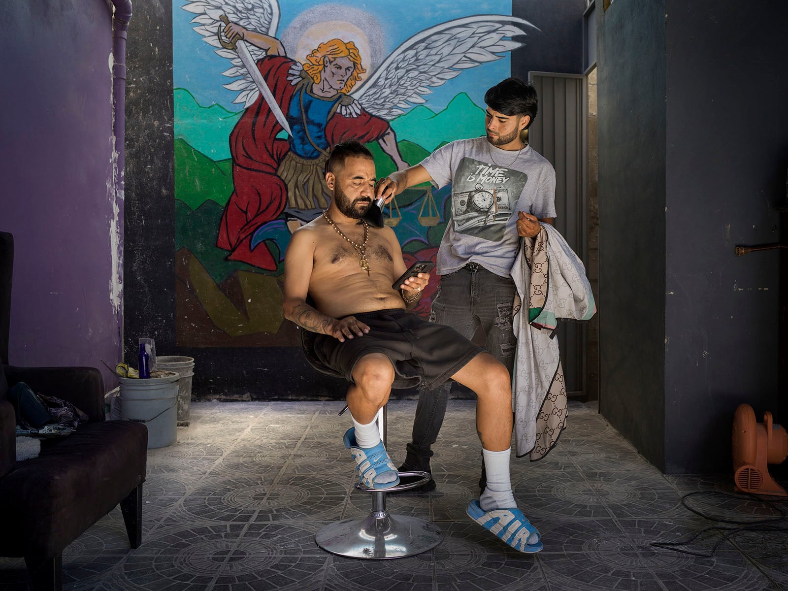 A man sits shirtless on a stool, looking at his phone, while another man stands behind him holding a barber cape. A colorful mural of an angel with a sword is painted on the wall in the background.