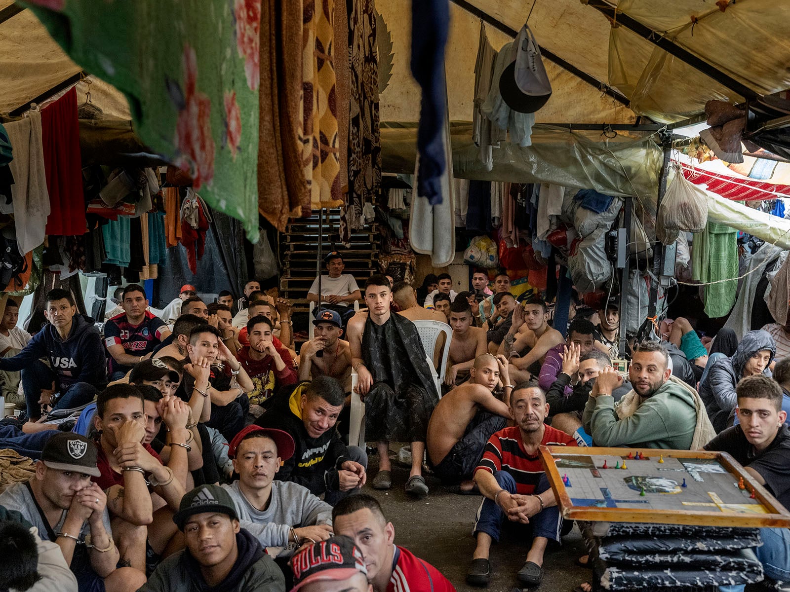 A large group of men sit closely together inside a crowded tent, surrounded by hanging blankets and clothes. Some look at the camera while others gaze elsewhere, creating a somber and contemplative atmosphere.