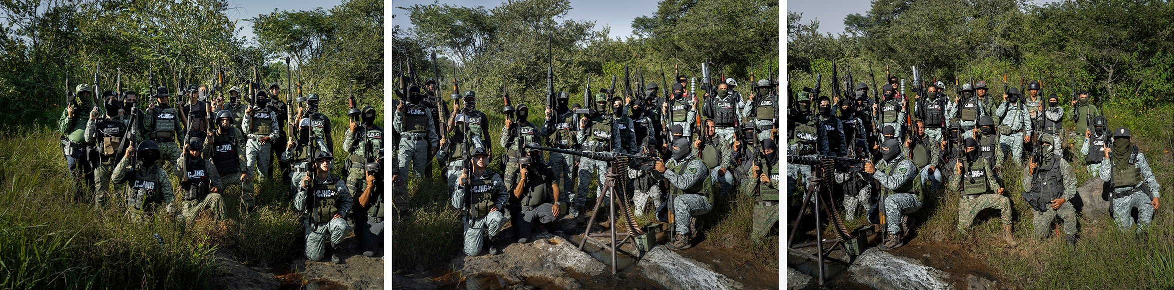 A large group of armed soldiers in full uniform and tactical gear poses together outdoors in a grassy, wooded area under a clear sky.