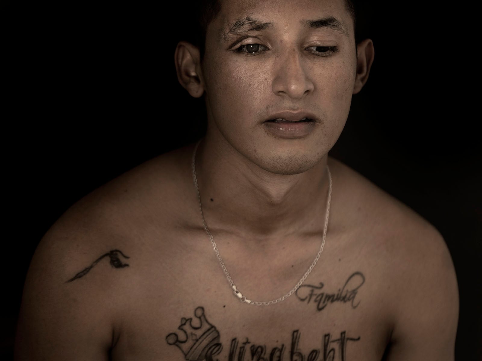 A young man with short hair, wearing a thin chain necklace, looks down with a neutral expression. He has facial scars and several tattoos on his chest, including the words "Familia" and "Elizabeth." The background is dark.