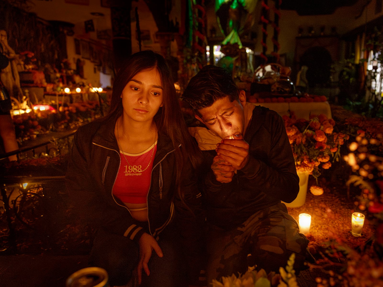 A young woman and a young man sit surrounded by candles and marigold flowers at night. The woman looks thoughtful, while the man lights a cigarette. The scene is warm and illuminated by candlelight.