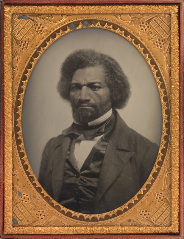 A black-and-white portrait of a stern-faced man with natural hair, wearing a suit and bow tie, set in an ornate gold oval frame with intricate designs.