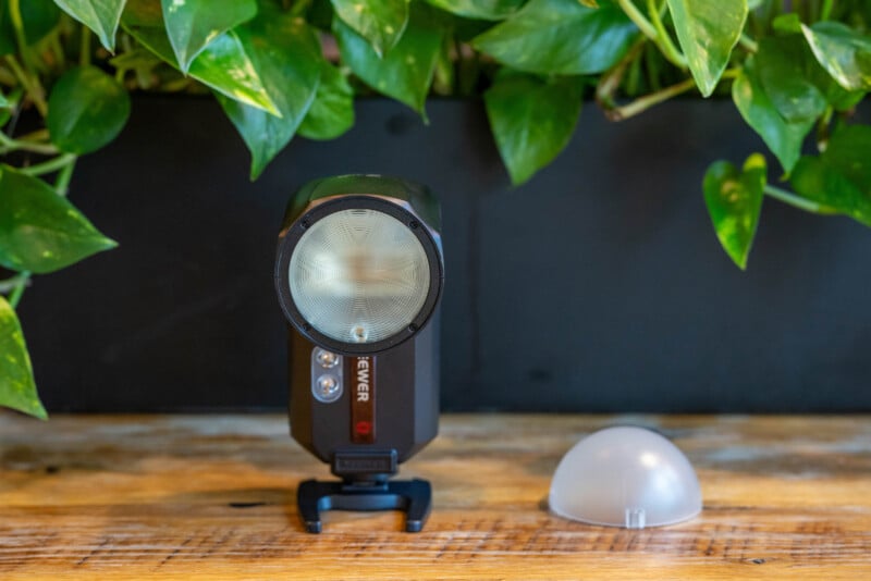 A camera flash unit with two buttons is standing on a wooden surface, in front of a leafy green plant. A translucent dome diffuser is placed to the right of the flash.