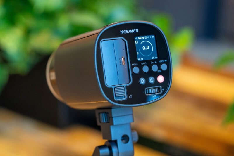 A close-up of a Neewer photography lighting device on a stand, showing its control panel with buttons, display screen, and adjustment settings. The background is blurred with green foliage and wooden surfaces.