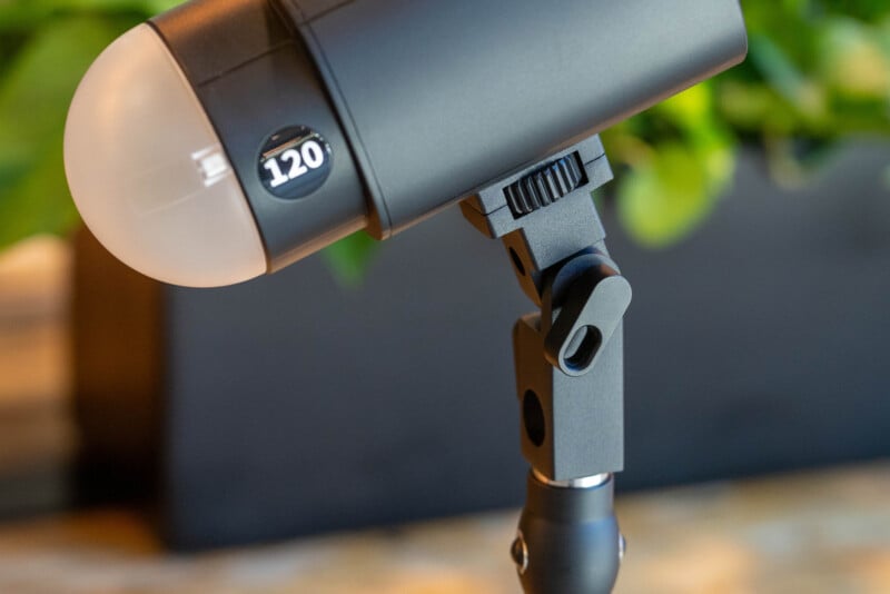 A close-up of a black studio light or flash unit labeled "120" on an adjustable stand, with a blurred green background.