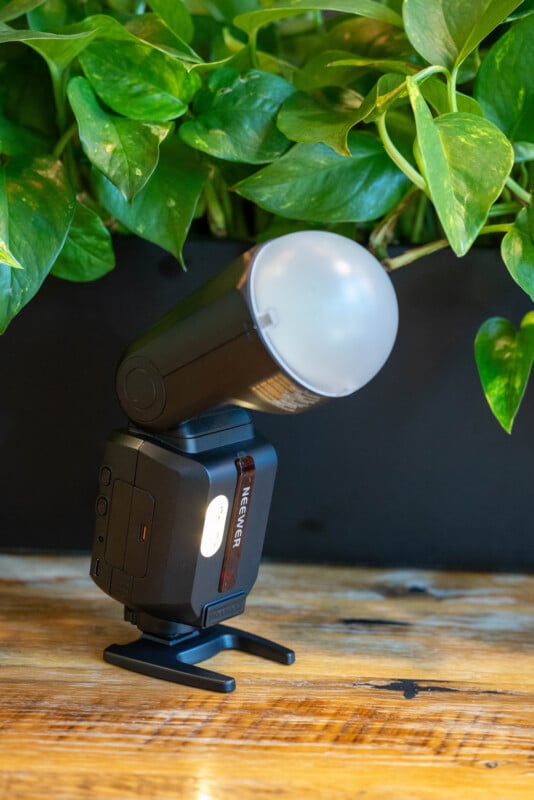 A black Neewer camera flash with a diffuser is placed on a wooden surface, with green leafy plants in the background.