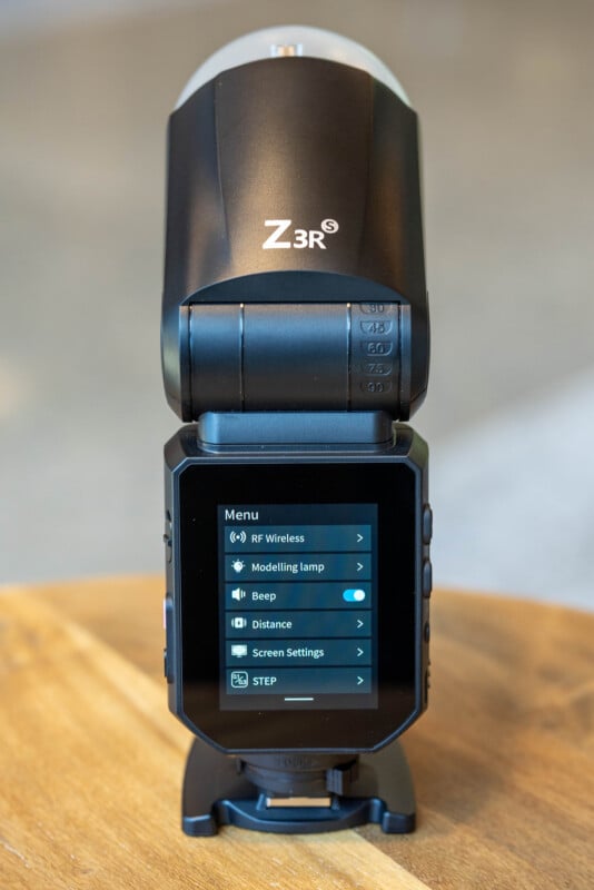 A close-up of a black Z3R camera flash unit with its digital menu screen displaying options such as RF Wireless, Modelling lamp, Beep, Distance, and Screen Settings, sitting on a wooden surface.