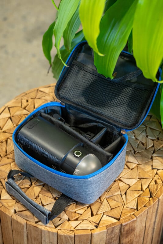 A small, open gray carrying case with a blue trim containing a black electronic device sits on a wooden surface, with a green leafy plant overhead.