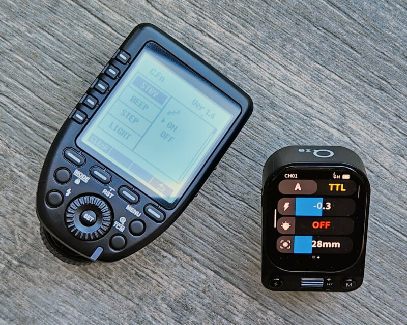 Two electronic camera accessories rest on a wooden surface: a handheld remote control with a display and buttons on the left, and a compact rectangular device with a color screen and icons on the right.