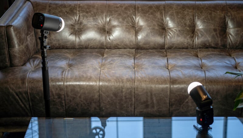 A brown, tufted leather couch sits behind a glass table, with two studio lights on tripods positioned in front of it, one on each side.
