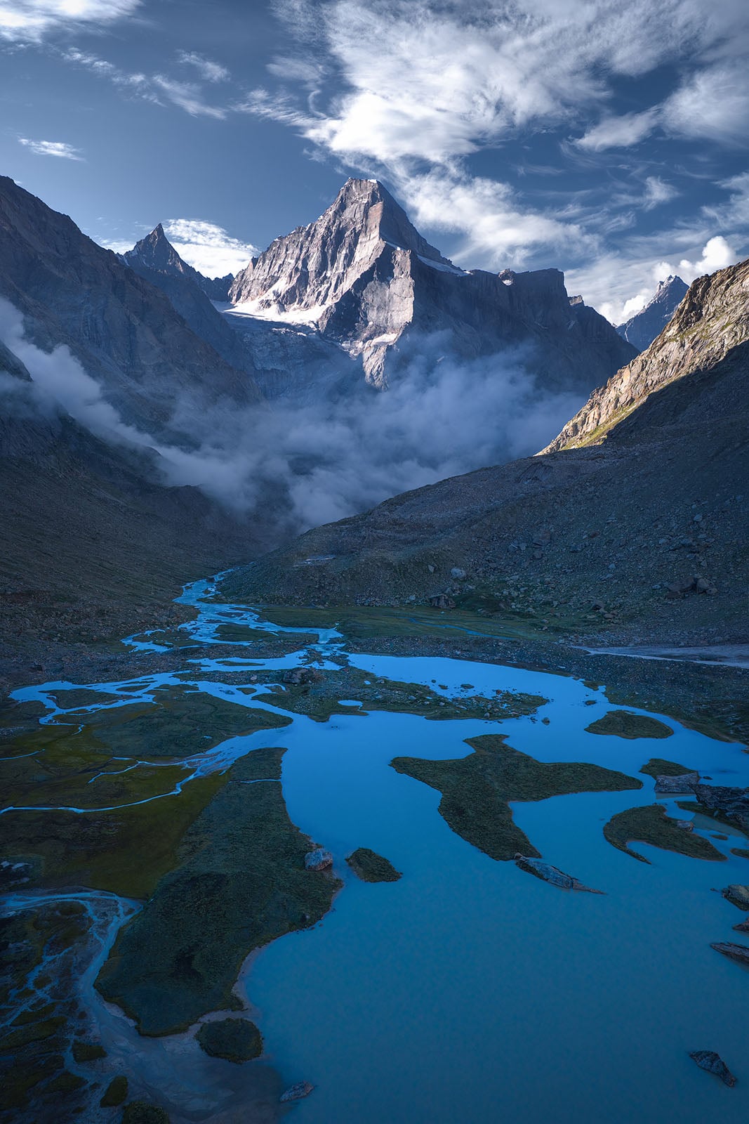 A winding blue river flows through a rocky valley with patches of green moss, leading towards towering, sunlit snow-capped mountains beneath a dramatic sky with clouds. Mist rises near the mountain base.