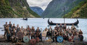 A group of people dressed in Viking-style costumes pose with shields and weapons by a fjord, with wooden Viking ships on the water and steep, green mountains in the background.