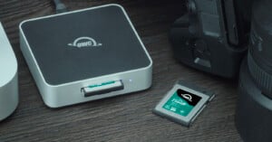 A close-up of an OWC memory card reader on a wooden surface, with a 256GB OWC memory card partially inserted and another card next to a camera and a lens. The card reader has a glowing indicator light.